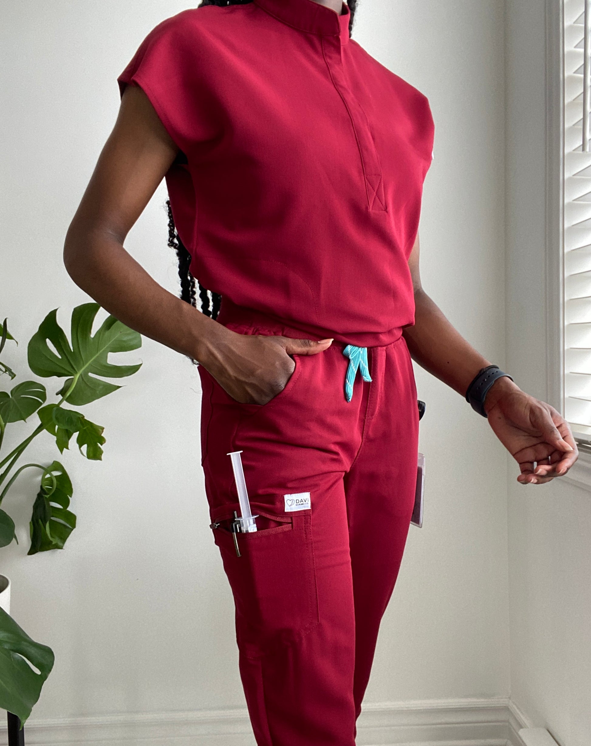 Black woman weary burgundy nursing scrub close to a window and a plant