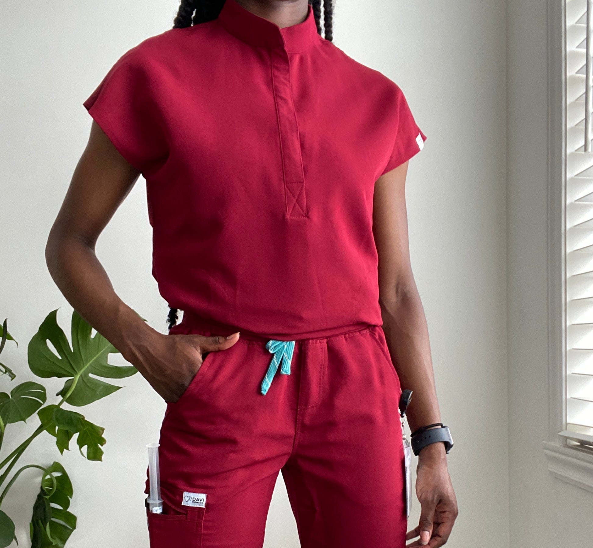 Black woman weary burgundy nursing scrub close to a window and a plant